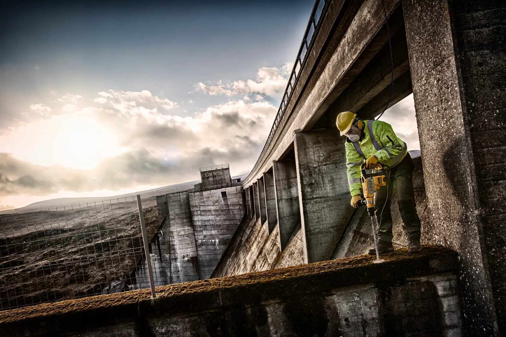 Trabajador usa taladro DEWALT en estructura de concreto al aire libre al atardecer, con equipo de seguridad.