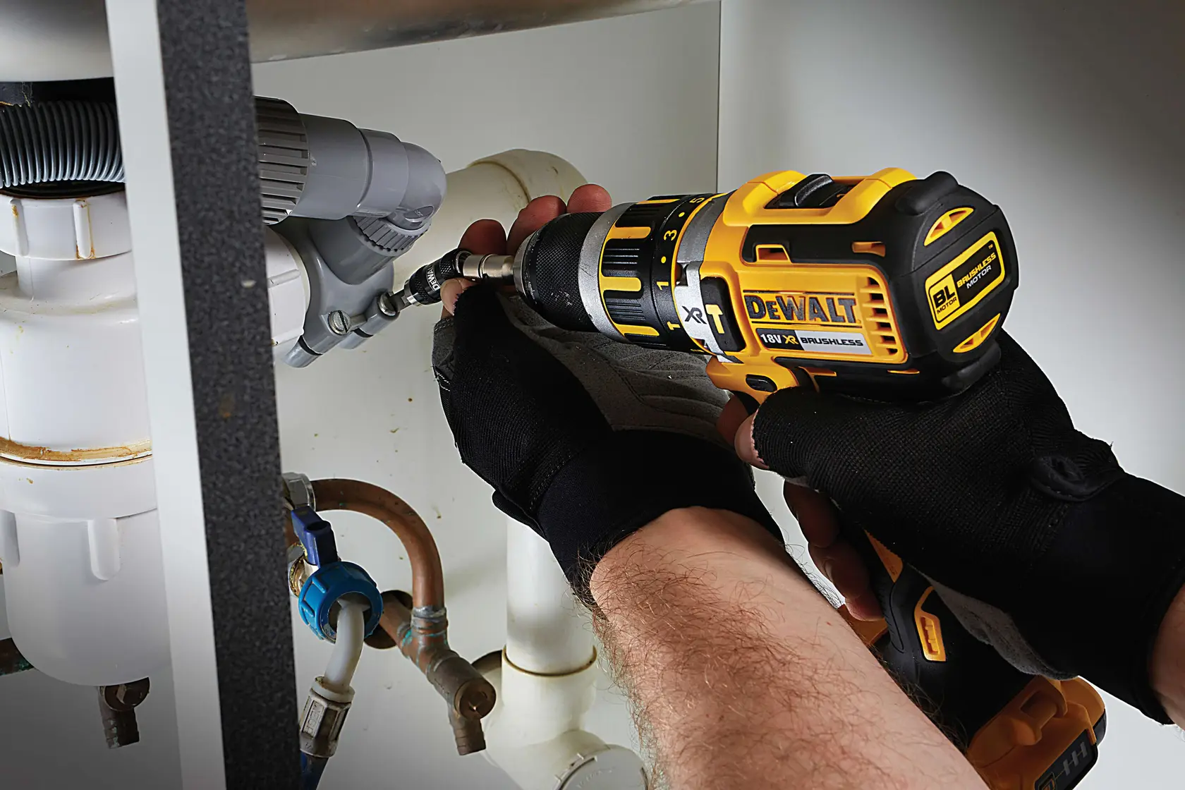 Foto de taladro DEWALT DT70518T usado por manos enguantadas en plomería bajo un fregadero.