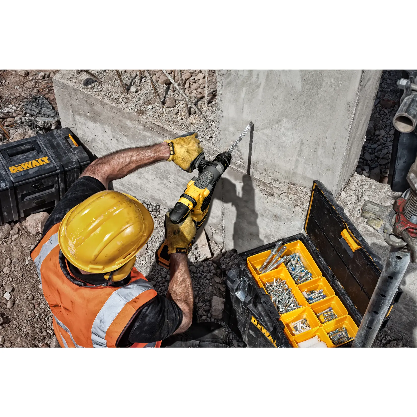 Trabajador perfora concreto con herramienta DeWalt, rodeado de cajas y sujetadores en obra.