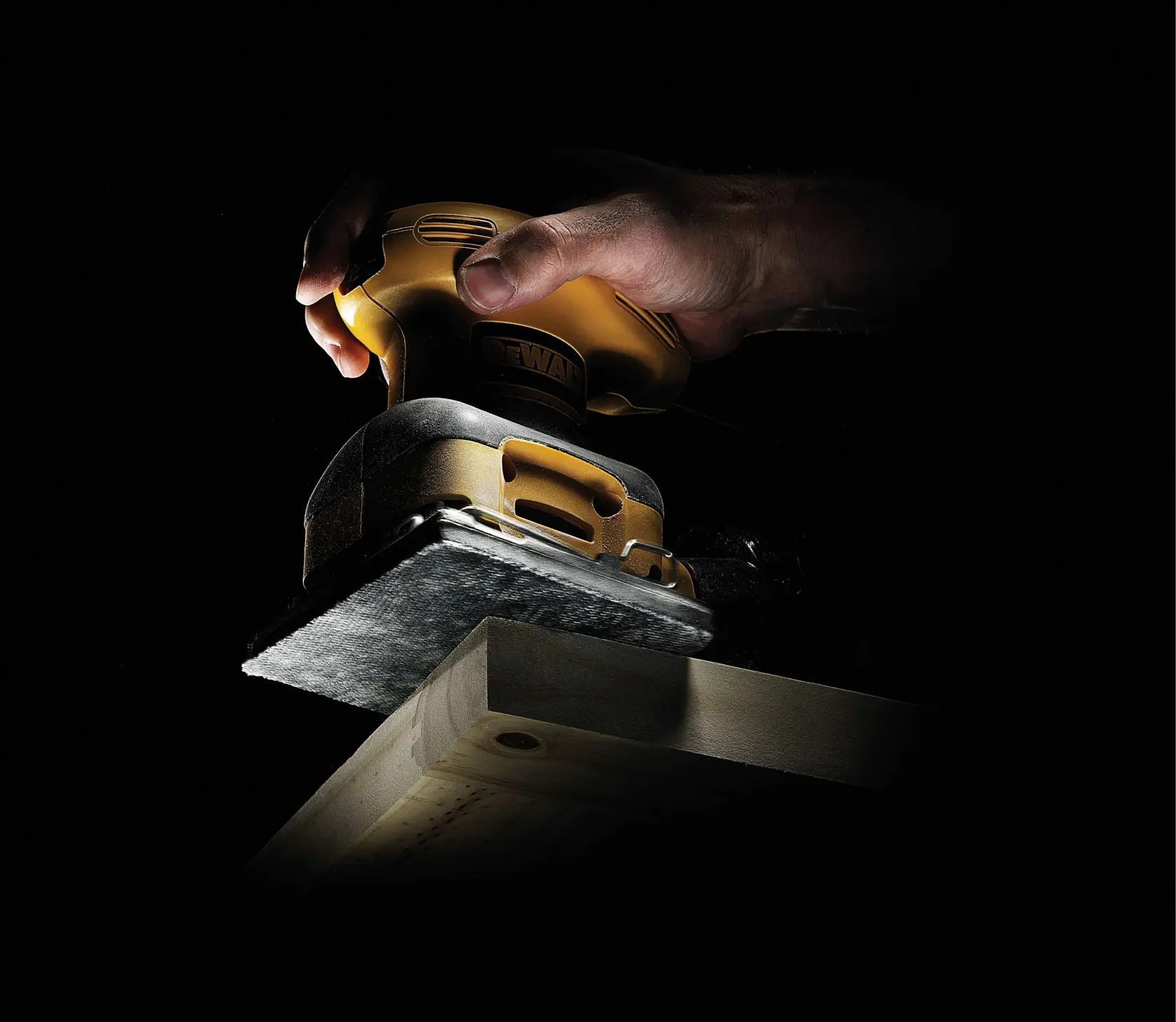 Mano usando una lijadora DEWALT para suavizar el borde de una tabla de madera con fondo oscuro.