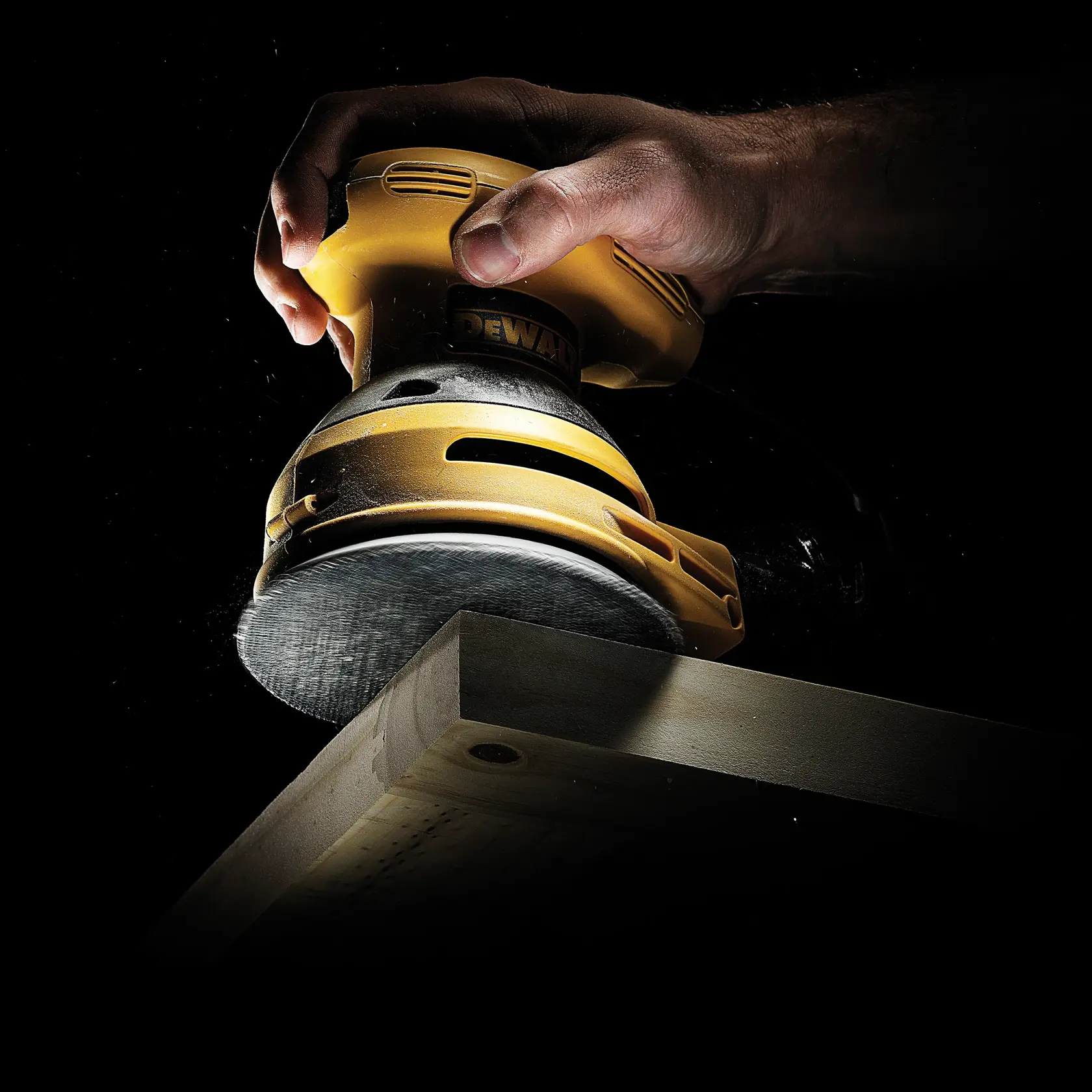 Mano con lijadora DEWALT suaviza el borde de una tabla de madera sobre fondo oscuro.