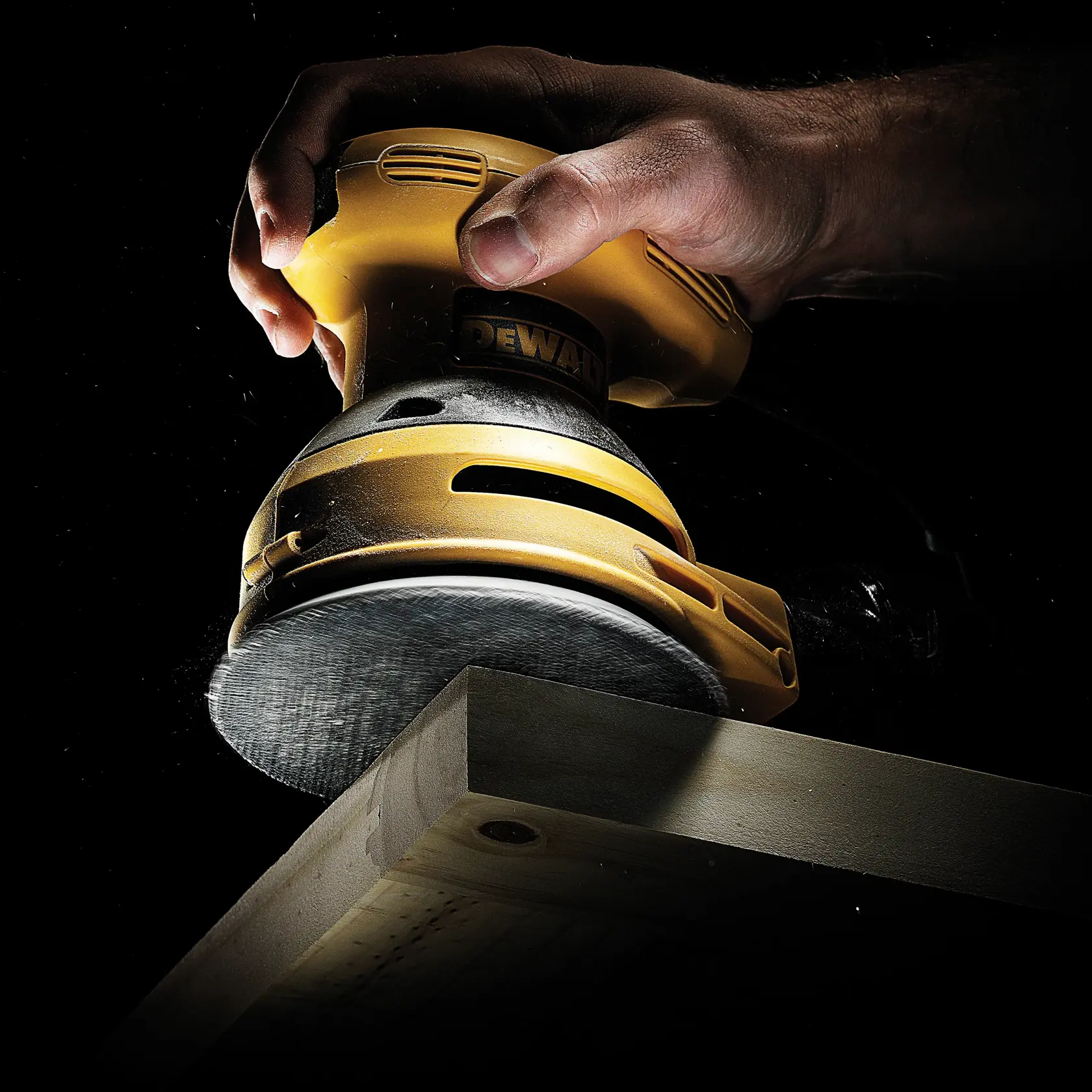 Mano sujetando una lijadora DEWALT, suavizando una tabla de madera sobre fondo oscuro.
