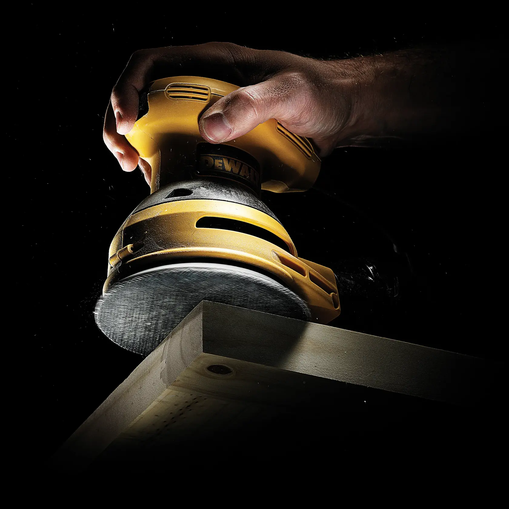 Mano usando lijadora DEWALT para suavizar el borde de una tabla de madera sobre fondo negro.