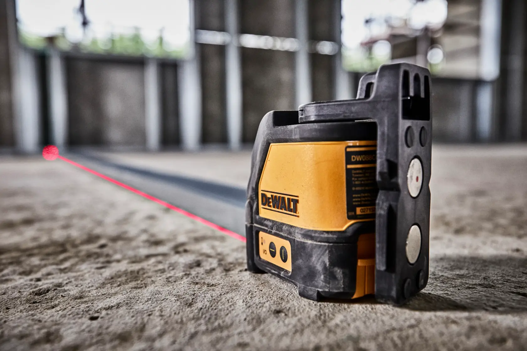 A DEWALT DW088K laser level is mounted on a metal column, projecting a red horizontal line across a construction site interior. In the background, a worker wearing a safety vest and helmet is aligning materials along the laser line.
