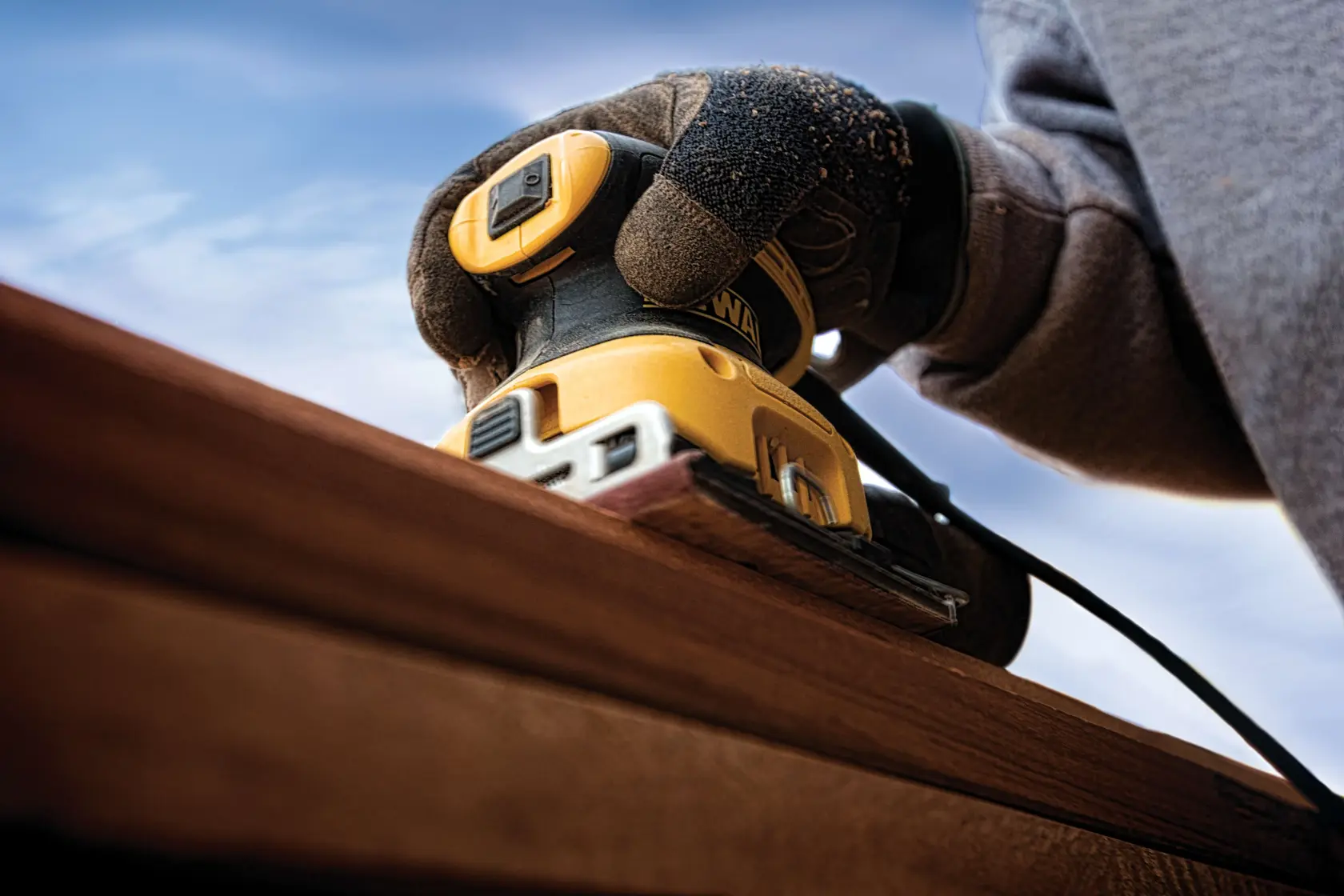 Primer plano de una persona usando una lijadora eléctrica DEWALT DWE6411 amarilla y negra para suavizar una superficie de madera al aire libre. La persona lleva guantes protectores.