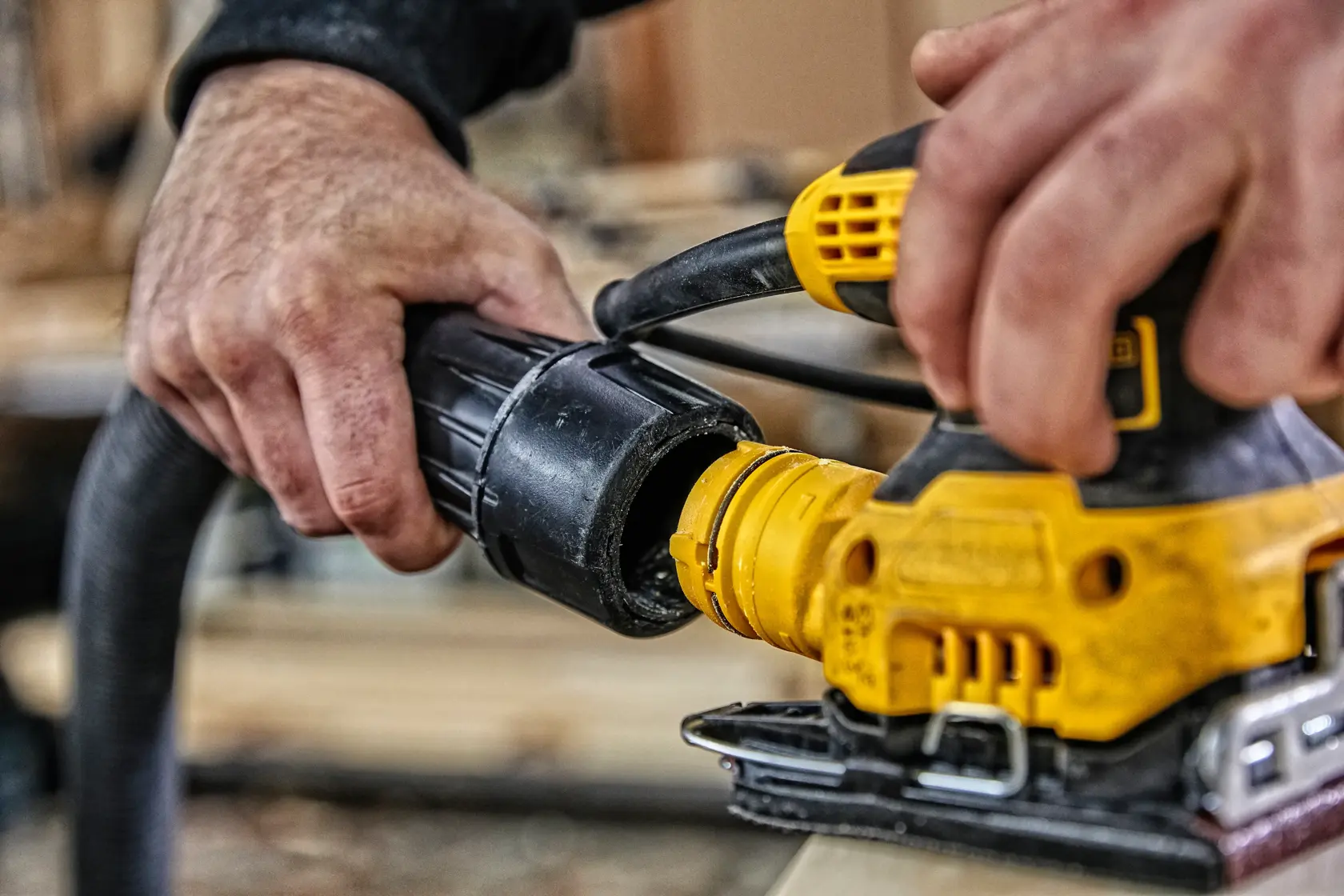 Una persona usando una lijadora eléctrica DEWALT sobre una superficie de madera dentro de un taller o garaje. La lijadora es amarilla y negra, y la mano de la persona la sostiene contra la madera.