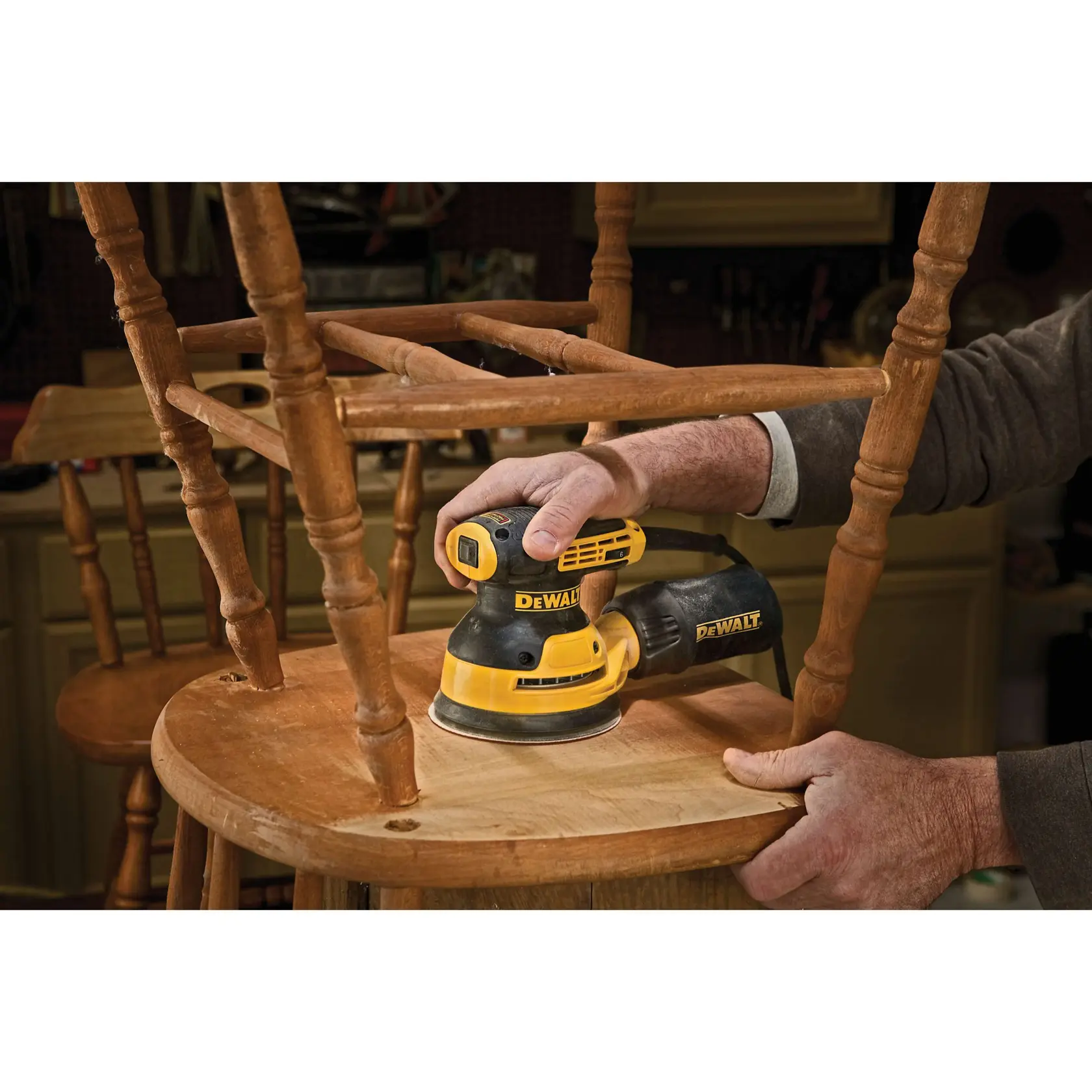 Une personne utilise une ponceuse orbitale aléatoire DEWALT DWE6423 pour poncer une porte en bois dans un atelier de menuiserie. Des pièces métalliques sont placées sur la table à proximité, et la personne porte un masque de protection.