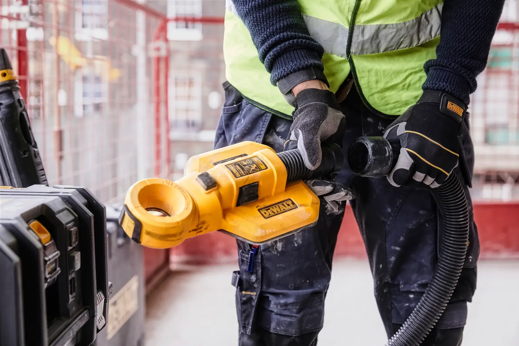Un travailleur portant un gilet de sécurité vert et un casque utilise un système d'extraction de poussière DEWALT DWH050 fixé à une perceuse jaune, perçant dans un plafond en béton dans un environnement industriel.