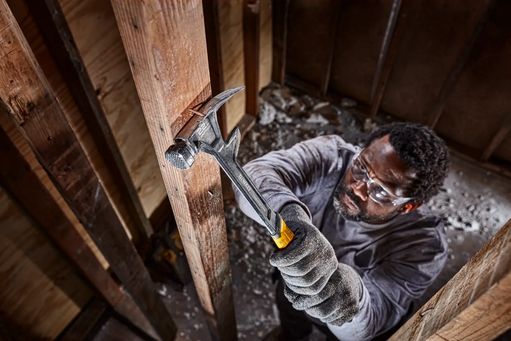 Una persona con guantes utiliza un martillo DEWALT con mango amarillo y negro para golpear una viga de madera dentro de un edificio en construcción. La cara de la persona está desenfocada.