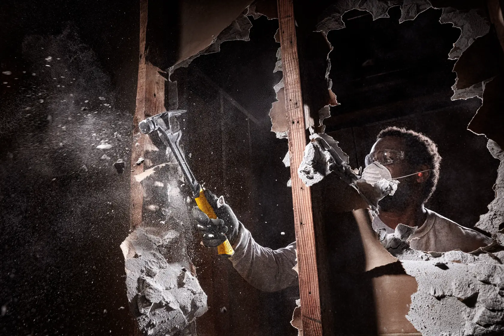 Una persona con guantes utiliza un martillo DEWALT para demoler una pared de yeso, creando una nube de polvo y escombros.