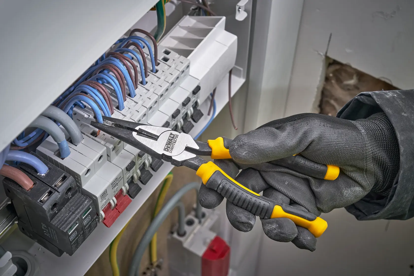 Mano enguantada usa alicates DEWALT para sujetar cables en panel de interruptores.