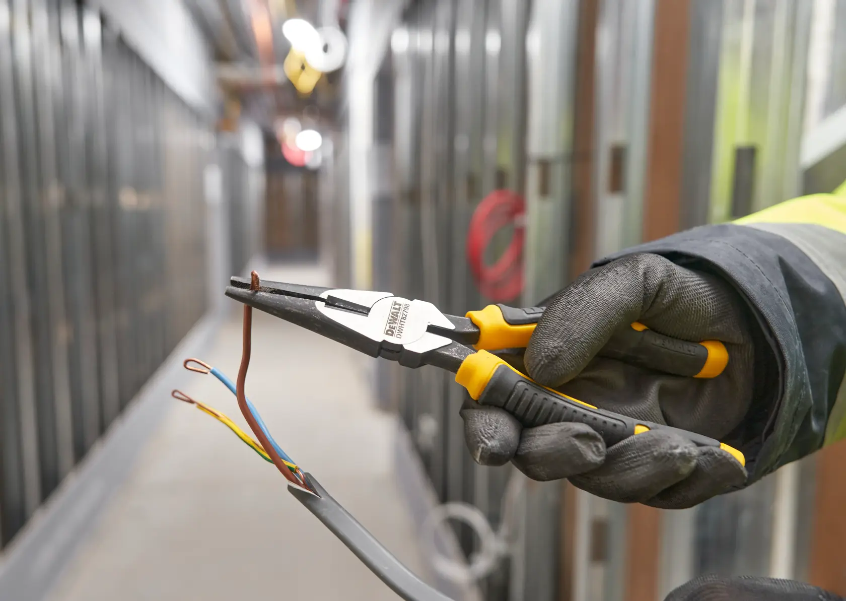 Mano enguantada usa alicates DEWALT para sujetar cables en un pasillo industrial.