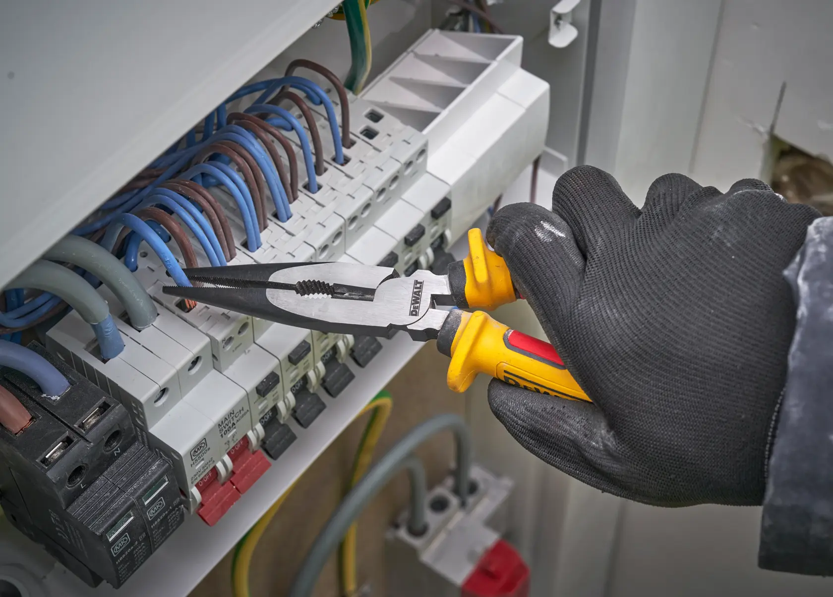 Mano enguantada usa alicates DEWALT para manipular cables eléctricos en un panel de interruptores.
