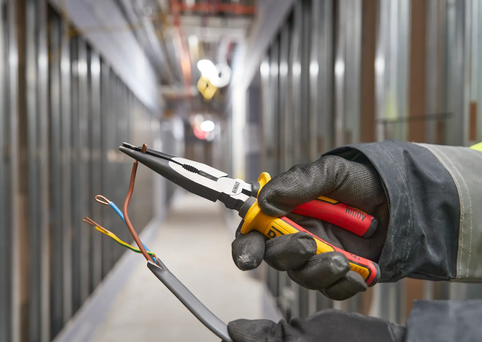 Mano con guante usa alicates DEWALT para pelar cables en un pasillo industrial.