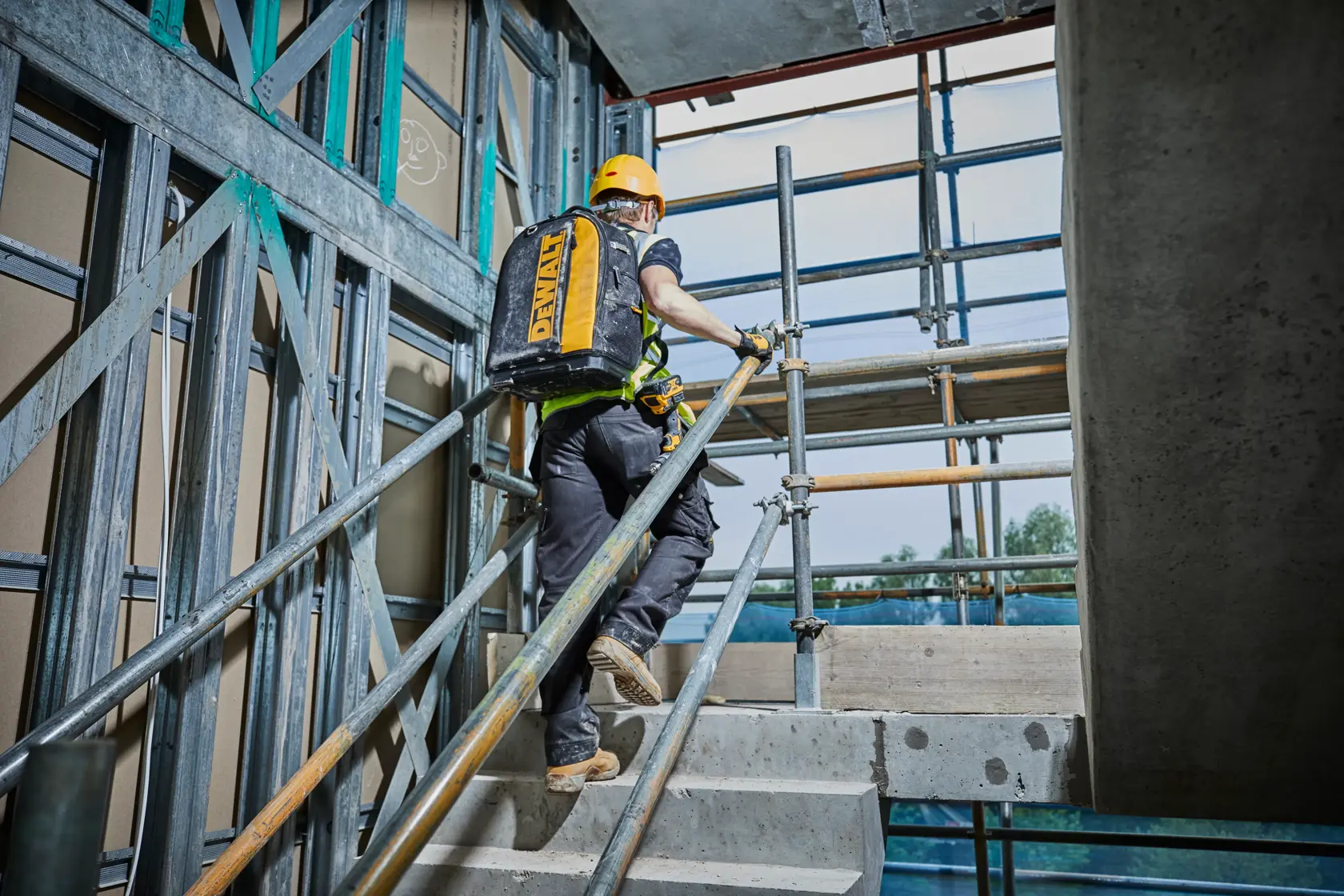 Trabajador sube escaleras en obra llevando una mochila amarilla DEWALT.
