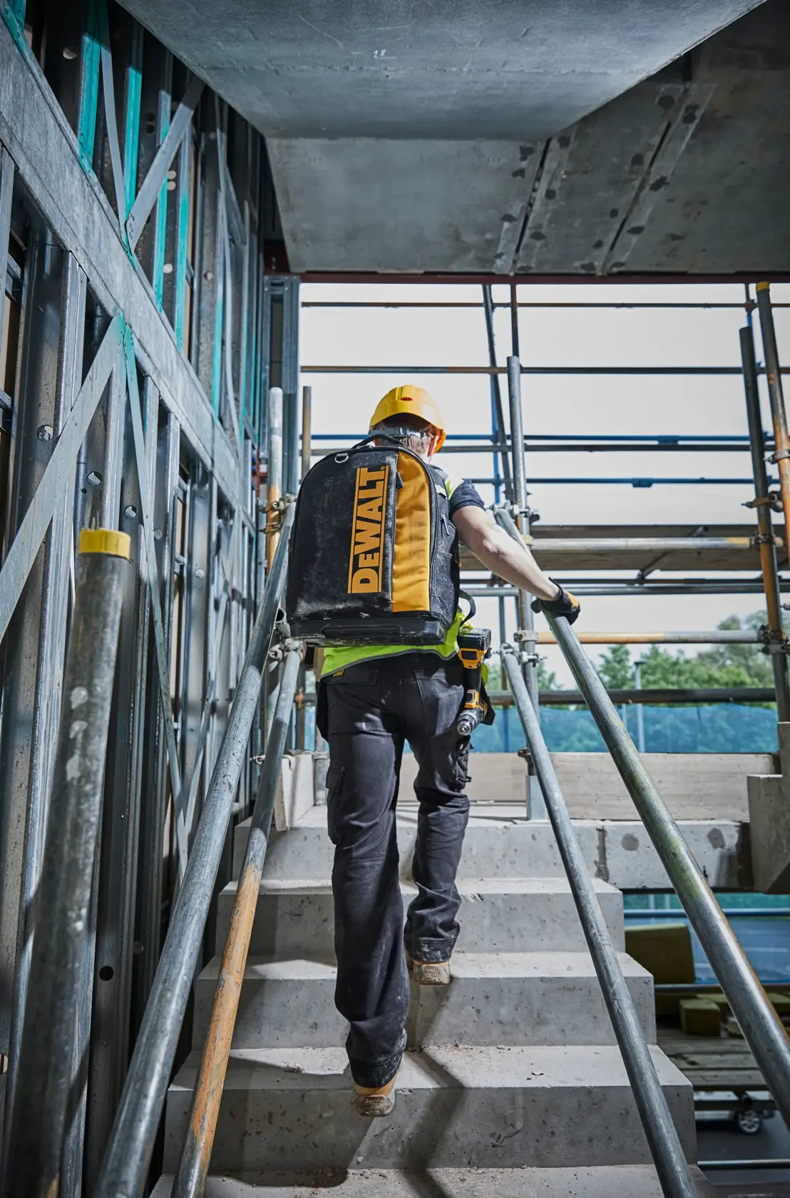 Trabajador con mochila DEWALT sube escaleras en obra, lleva casco amarillo y equipo de seguridad.