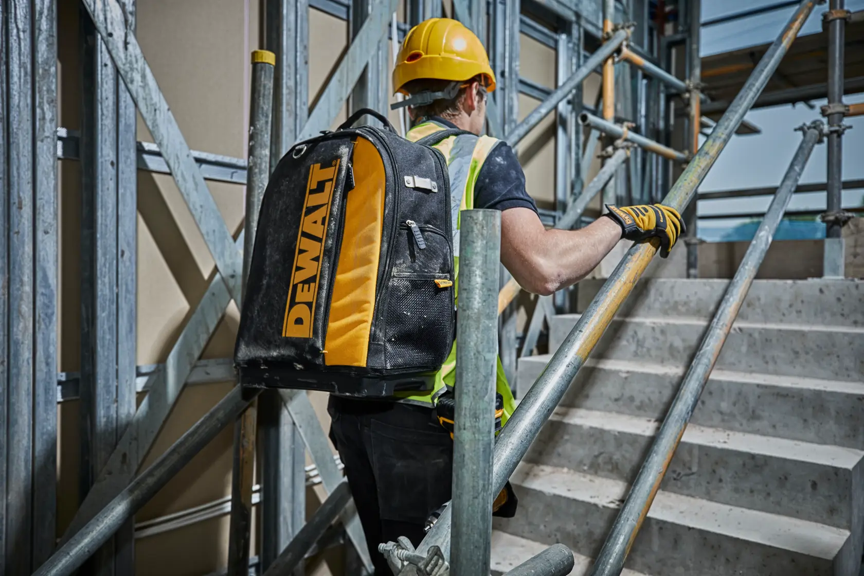 Trabajador con mochila DEWALT sube escaleras en obra, llevando casco y equipo de seguridad.