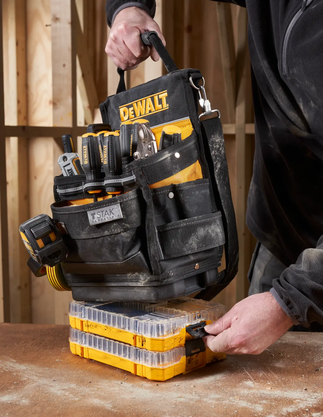 Bolsa de herramientas DEWALT con herramientas sobre organizadores amarillos, sostenida por persona con ropa de trabajo.