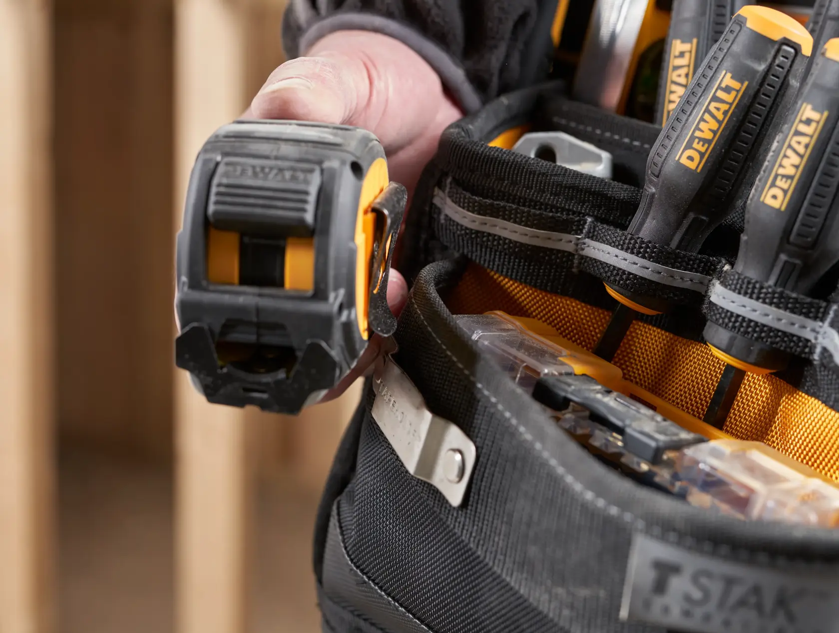 Primer plano de un cinturón DEWALT con mano sosteniendo cinta métrica y destornilladores en bolsillos.